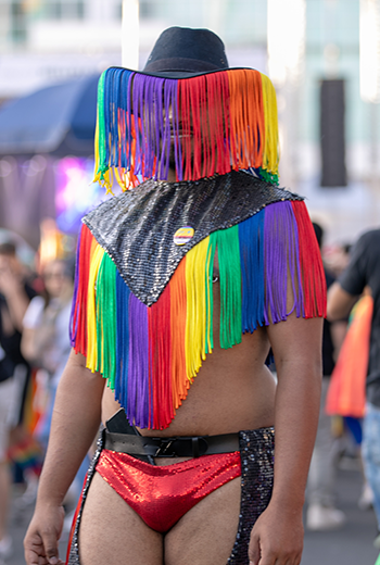 Foto de um homem gay com look personalizado na 7ª Marcha pela Diversidade de Curitiba, em 2024. Ele veste um manto preto e colorido, uma sunga preta e vermelha e um chapéu preto com linhas coloridas caindo da aba. Ele está posando para a foto e ao fundo vemos pessoas andando durante a marcha.