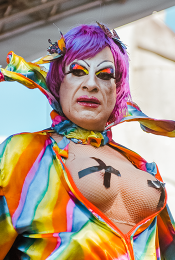 Foto da Drag Queen Tinna Simpson na 7ª Marcha pela Diversidade de Curitiba, em 2024. Ela veste uma blusa colorida e os peitos estão de fota com um "x" protegendo os mamilos. Ela olha para o horizonte e a foto captura um momento seu no palco