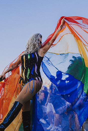 Foto de uma Drag Queen na 7ª Marcha pela Diversidade de Curitiba, em 2024. Ela veste um body preto com fitas coloridas e está usando uma capa nas cores da bandeira LGBTI+. Ela está movimentando a bandeira e a foto captura este momento.