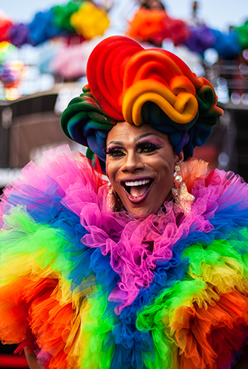 Foto da Drag Queen Yasmin Carraroh na 7ª Marcha pela Diversidade de Curitiba, em 2024. Ela veste um vestido todo colorido e feito de tule bufante e uma peruca de espuma toda colorida em formato de ondas. Ela está sorrindo para a foto olhando para o horizonte.
