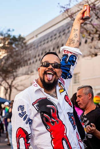 Foto de homem segurando uma bebida alcoólica para cima, sorrindo com a língua de fora, vestindo uma camiseta estampada e óculos de sol. Foto na 7ª Marcha pela Diversidade de Curitiba, em 2024