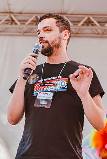 Foto do Diretor Lucas Siqueira, com o microfone na mão, falando no palco da 7ª Marcha pela Diversidade de Curitiba, em 2024